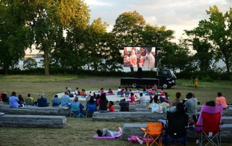 families and friends gather on a lawn and in lawn chairs to watch a movie on a mobile screen